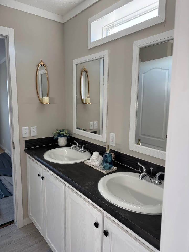 A modern bathroom with dual sinks set in a black countertop, white cabinets, and two mirrors. Elegant decor includes a gold-framed candle holder and small plant.