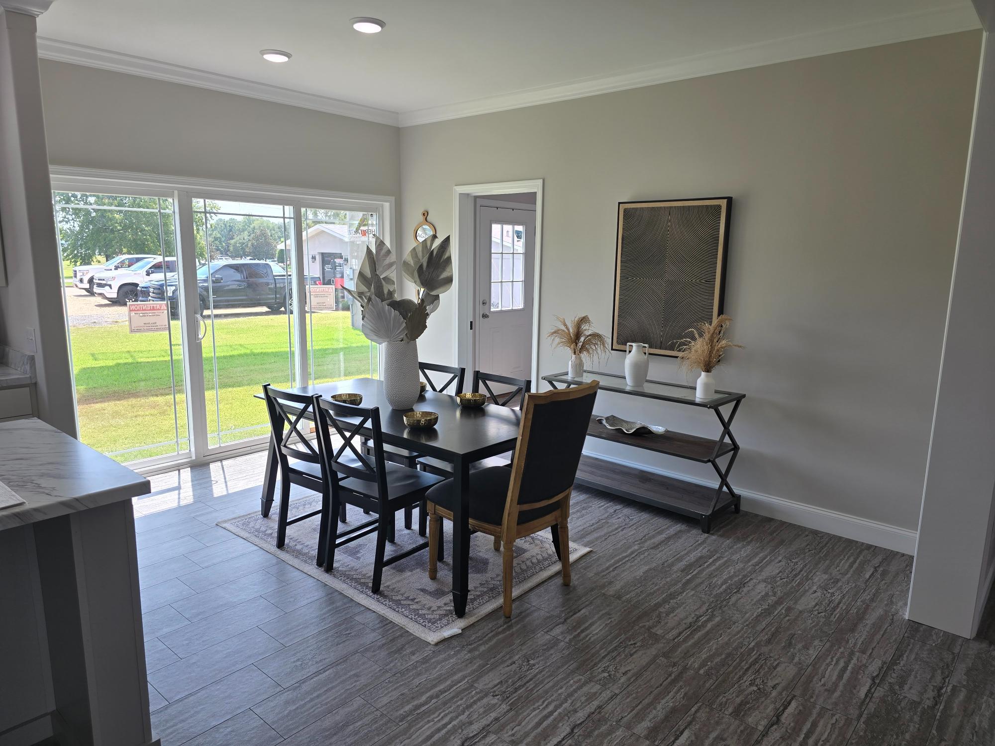 Modern dining room with a dark table set for six on a light rug. Large sliding glass doors show a grassy view. Neutral tones and decorative vases create a calm ambiance.
