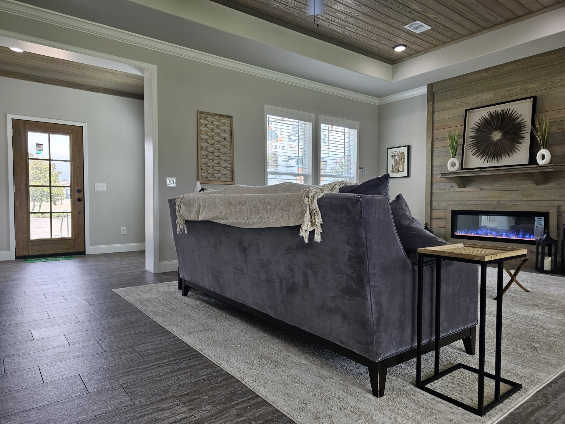 Modern living room with a gray couch, beige throw, and small side tables. A fireplace below decor on the wood-paneled wall adds warmth. Natural light from a door and windows enhances the cozy ambiance.