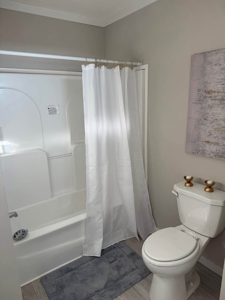 A small, modern bathroom with a white bathtub and shower curtain, a gray bath mat, a toilet with two gold ornaments, and neutral wall decor.