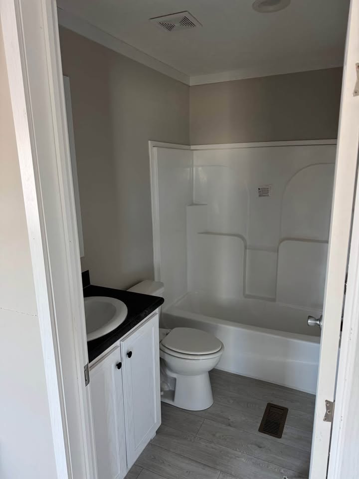 Small bathroom with a white bathtub and shower, white toilet, and a black countertop vanity with a sink. Light gray walls and wood-look flooring.