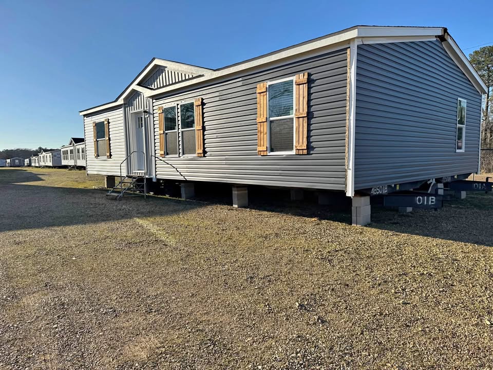 A gray modular home with wooden shutters sits elevated on a gravel lot under a clear blue sky. It exudes a simple, rustic charm.
