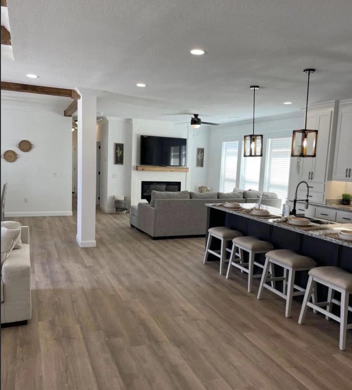 Spacious modern living area with wood floors, featuring a kitchen island with stools, a comfy gray sofa, large windows, and a wall-mounted TV above a fireplace.