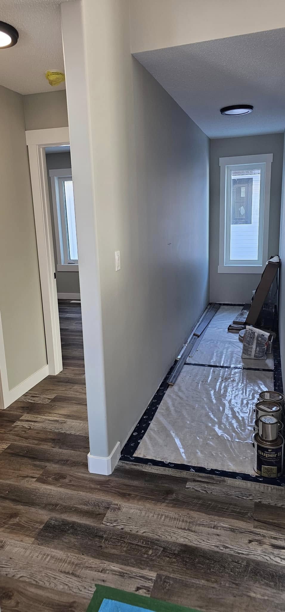 A hallway under renovation with gray walls, wood flooring, and paint cans on the right. Plastic sheeting covers part of the floor. Windows provide natural light.