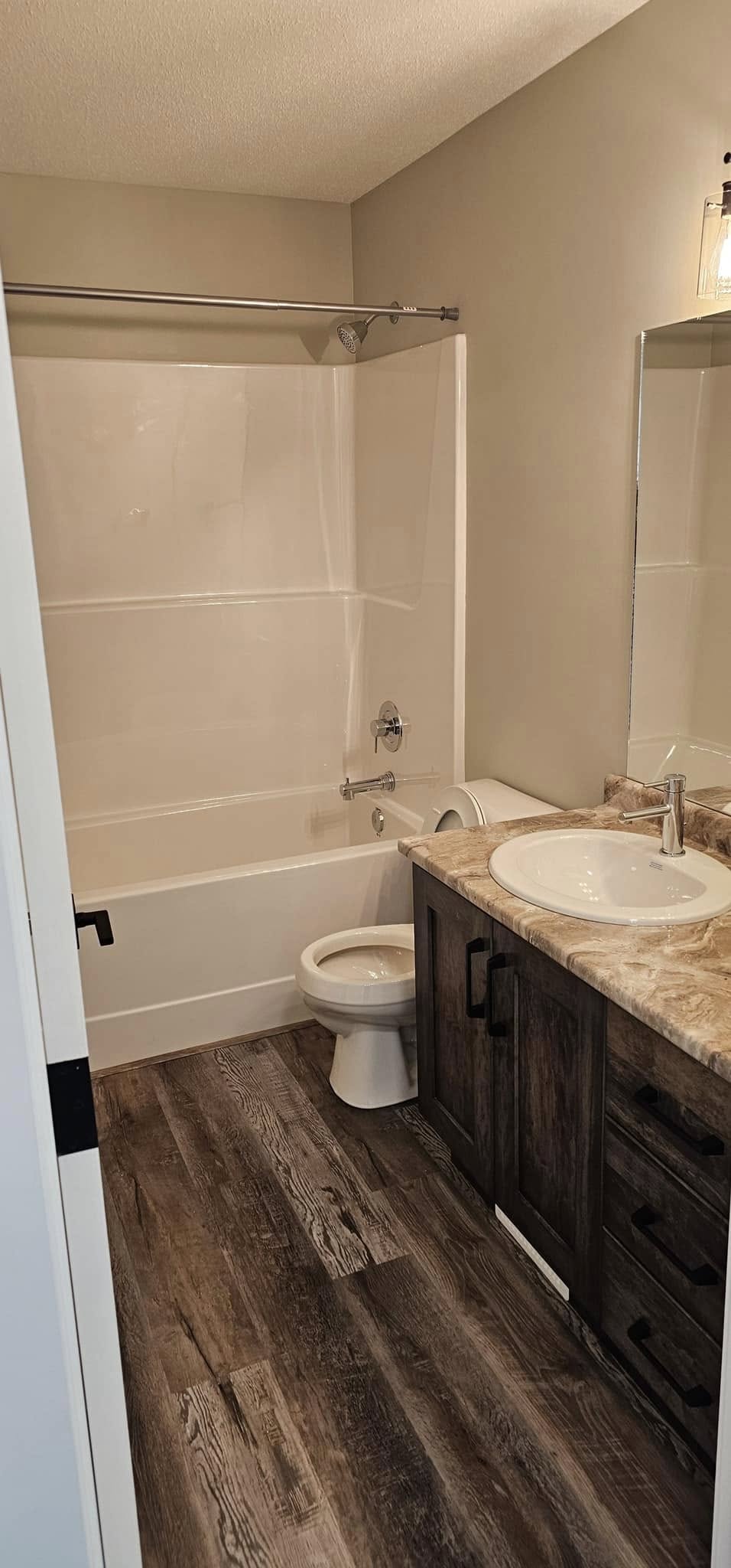 A bathroom with a white bathtub and shower curtain rod, a wooden vanity with a marbled countertop, a white sink, and a toilet. The floor is dark wood.
