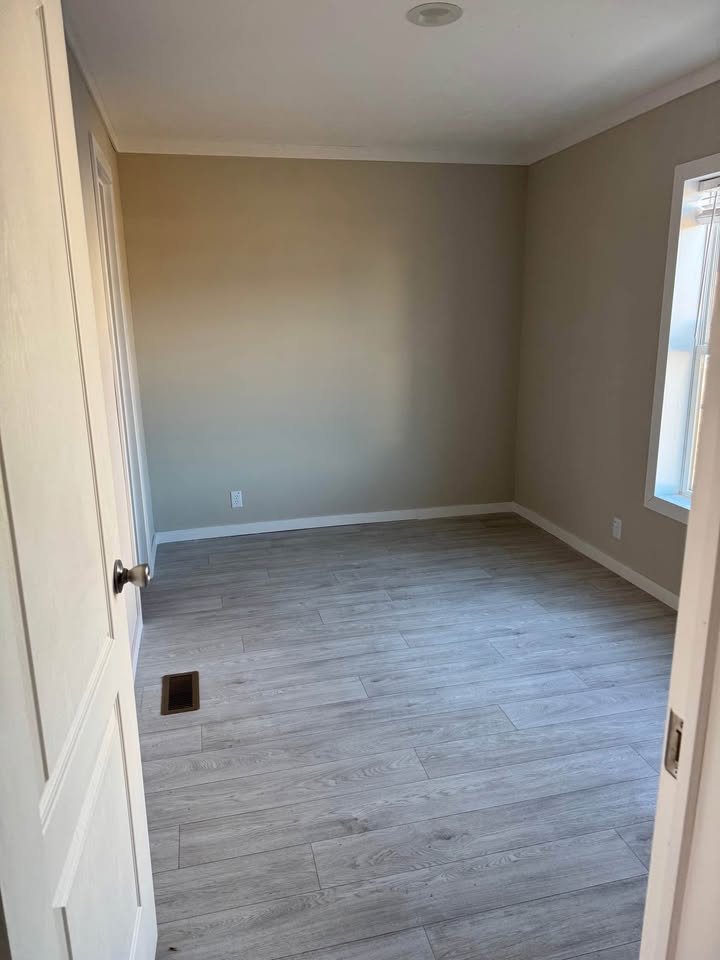Empty room with beige walls, light wood flooring, and a single window on the right. An open door is visible on the left. The atmosphere is calm and neutral.