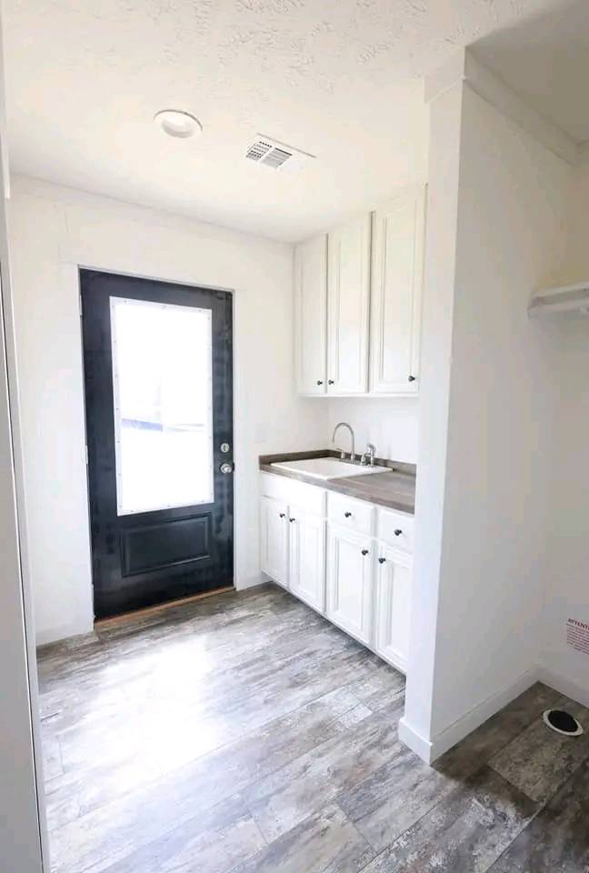 A bright laundry room with white walls and wooden flooring, featuring a black-framed door, cabinets, and a sink with a modern faucet, conveying a fresh, clean feel.