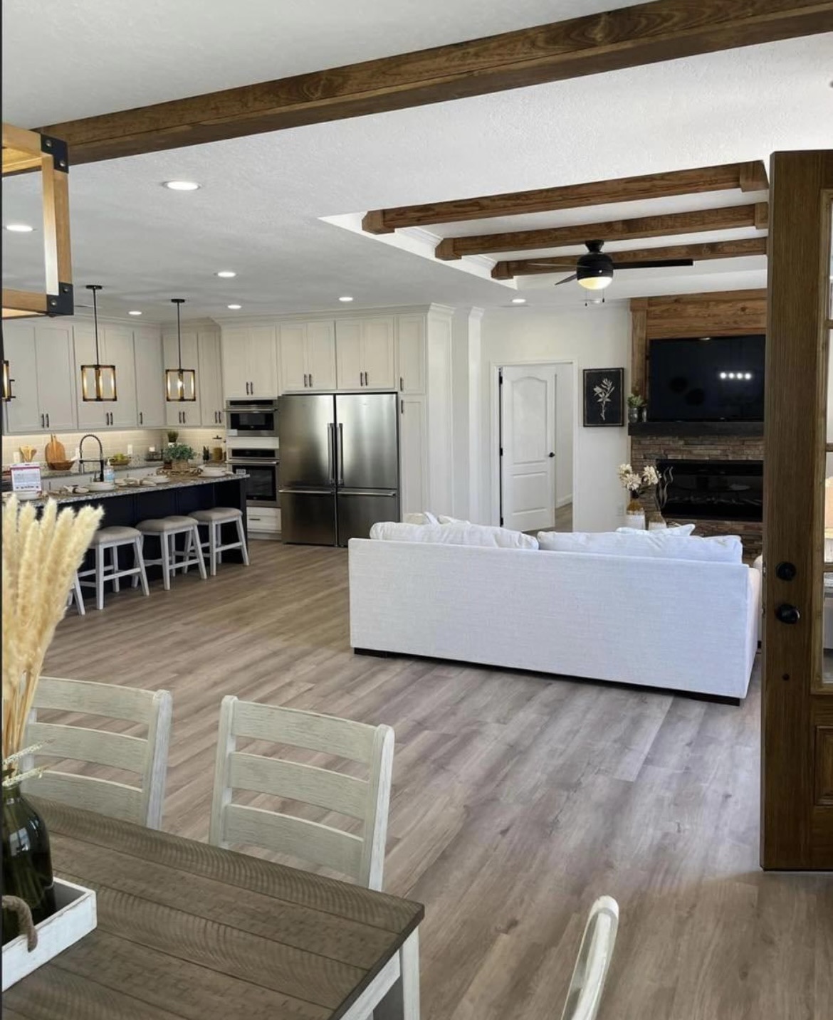 Spacious open-plan kitchen and living room with light wood floors, white walls, and dark wooden ceiling beams. Modern furnishings, calm ambiance.
