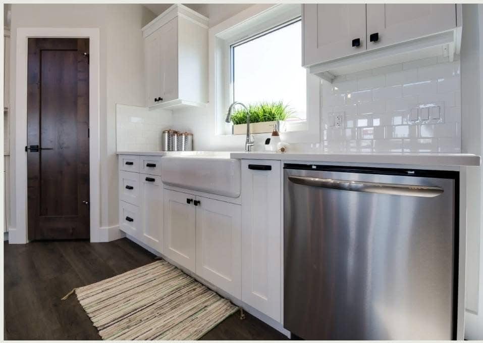 A modern kitchen with white cabinets, a farmhouse sink, and stainless steel dishwasher. A window above the sink shows greenery. A rug lies on dark wood floor.