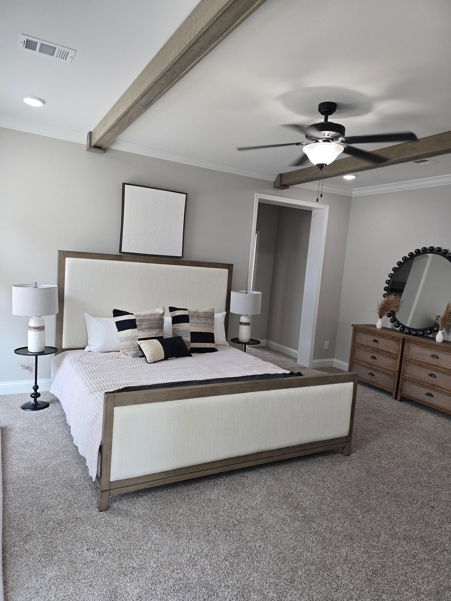 Spacious bedroom with a neutral palette. Features a large bed with patterned pillows, a ceiling fan, exposed beams, carpet, and a dresser with a round mirror. Calm ambiance.