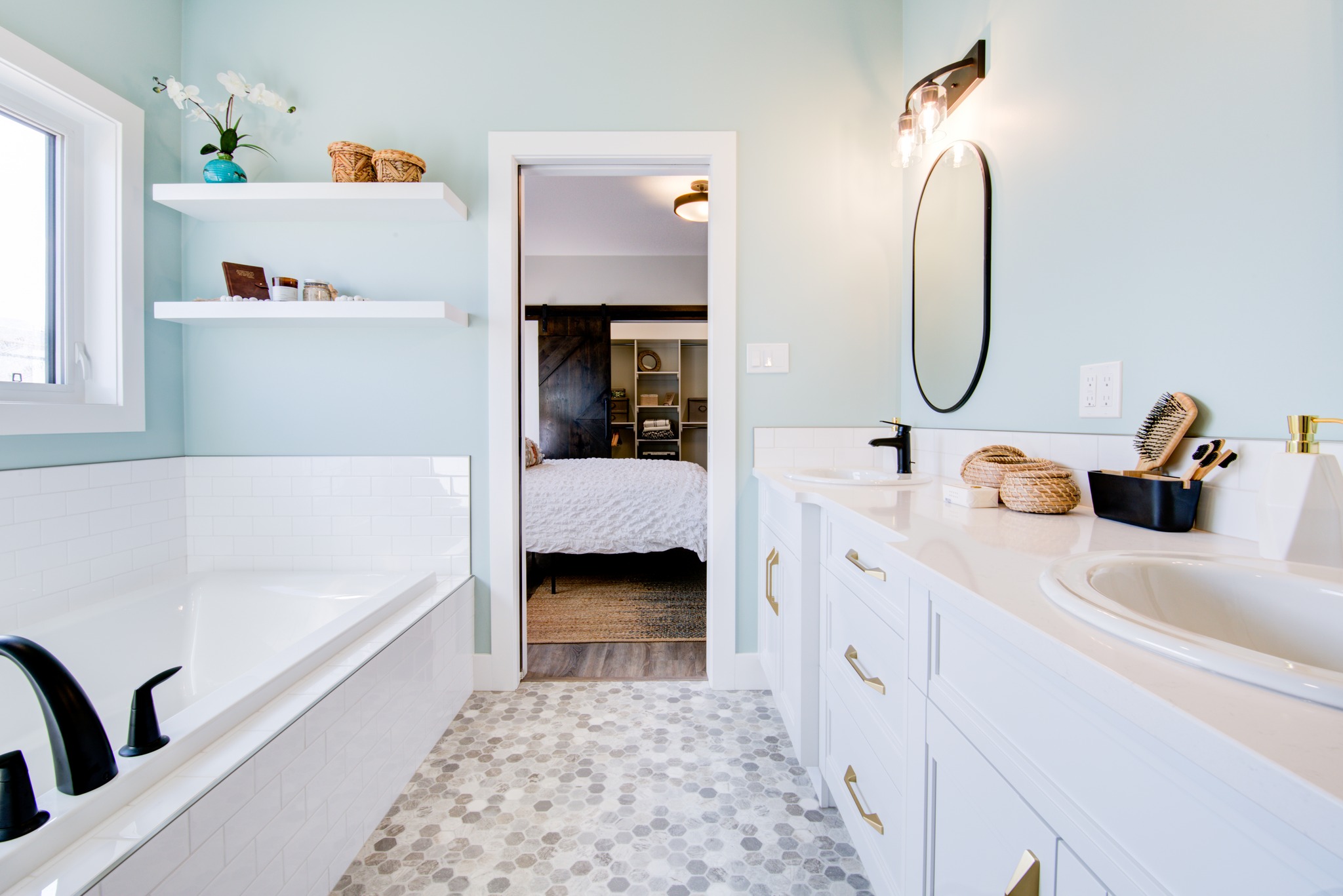 Bright bathroom with light blue walls, dual sinks, gold handles, a white tub, and circular mirror. Shelves hold decor; doorway shows a bedroom.