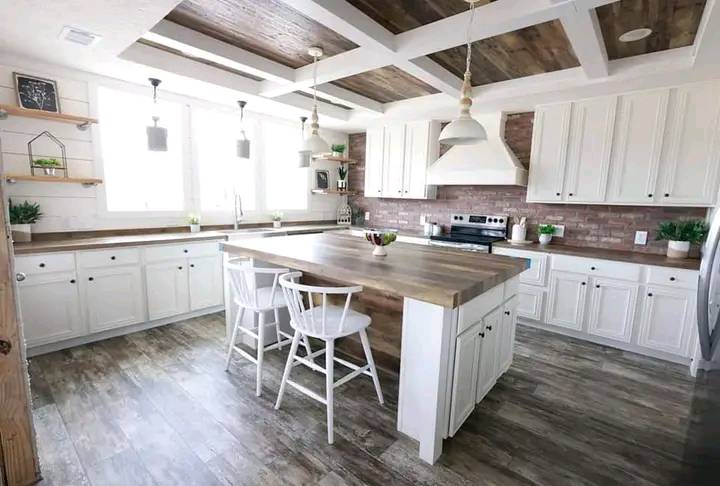 Spacious farmhouse kitchen with white cabinets, brick backsplash, and wooden island. Three pendant lights, two white chairs, and greenery accents. Cozy and inviting.