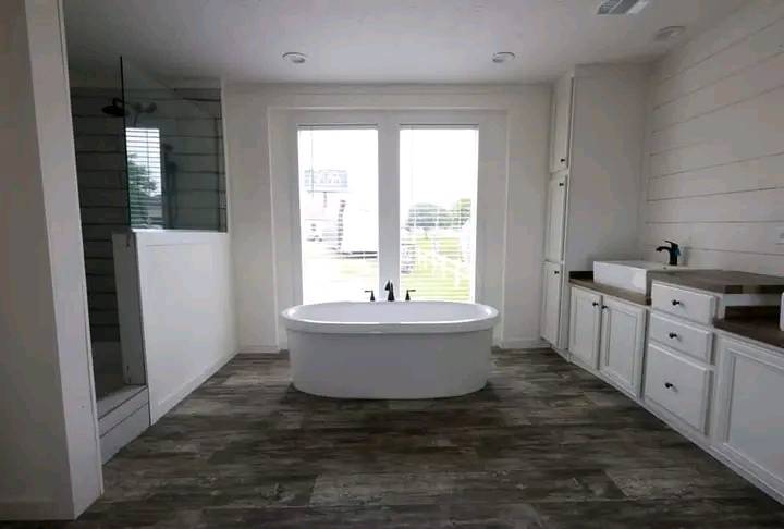 Modern bathroom with a freestanding bathtub centered under large windows, wooden flooring, a frameless glass shower on the left, and white cabinetry on the right.