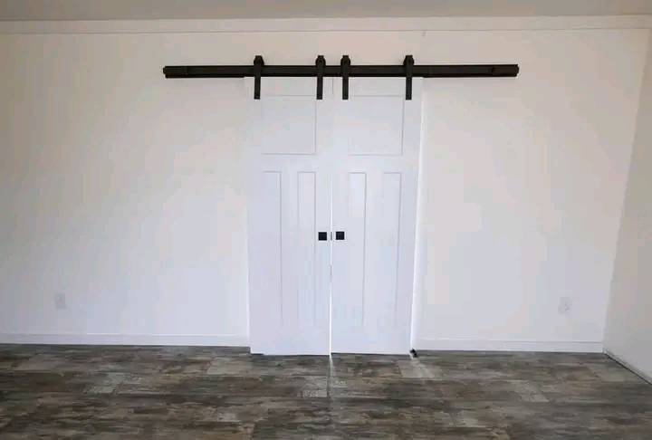 White sliding barn door on a track against a plain wall, with gray wood-patterned flooring. The scene is minimalistic and modern.
