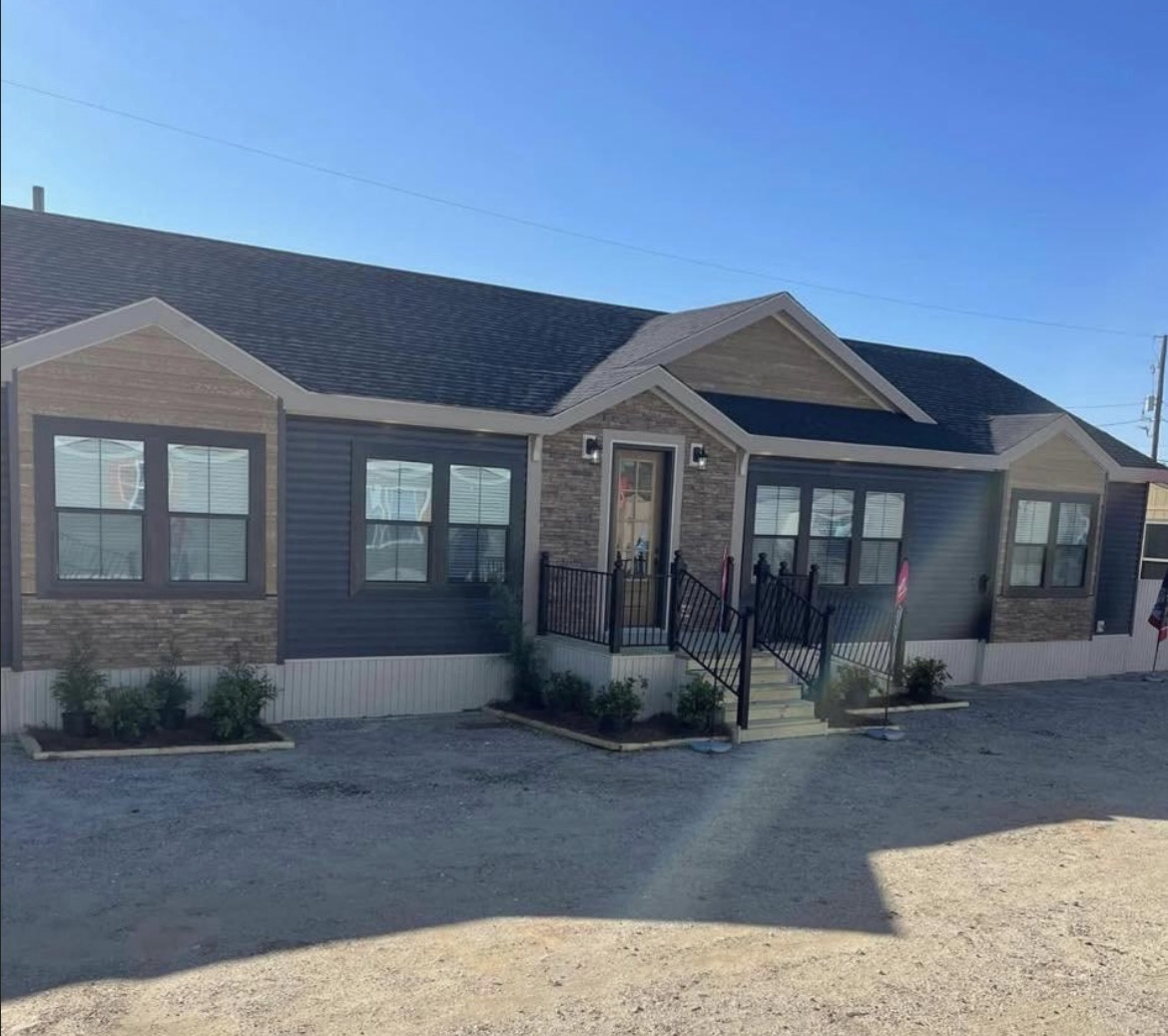A single-story modular home with a stone and dark siding exterior, featuring multiple windows and a central entrance. The setting is sunny, with a small dirt yard.