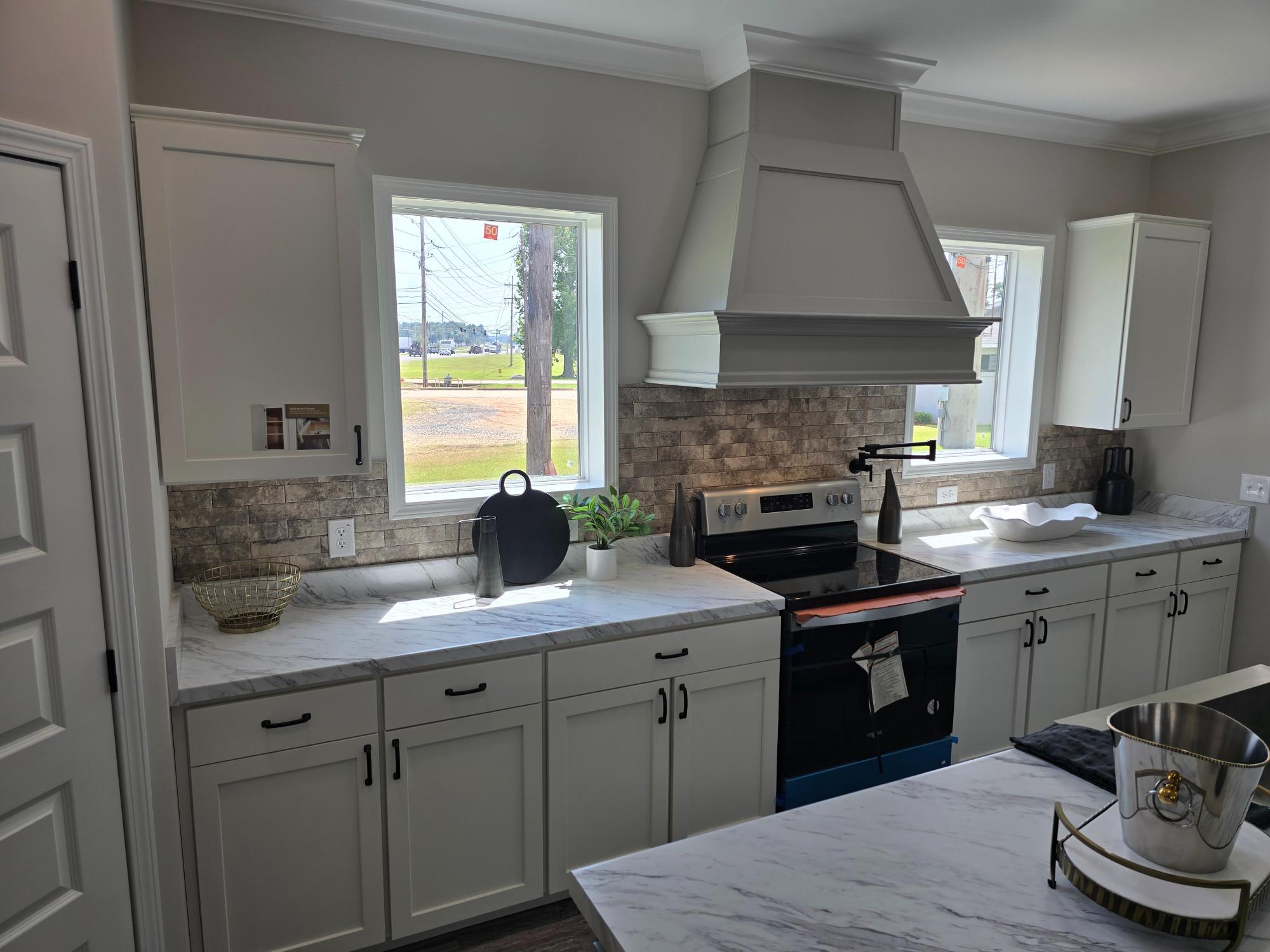Modern kitchen with white cabinets, marble countertops, and a stainless steel stove. Two large windows brighten the space. The room feels sleek and inviting.