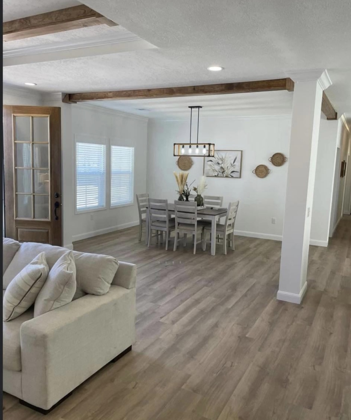 Bright living space with wooden floors featuring a cozy beige sofa, a dining table set for six under a modern chandelier, and wall art accents.