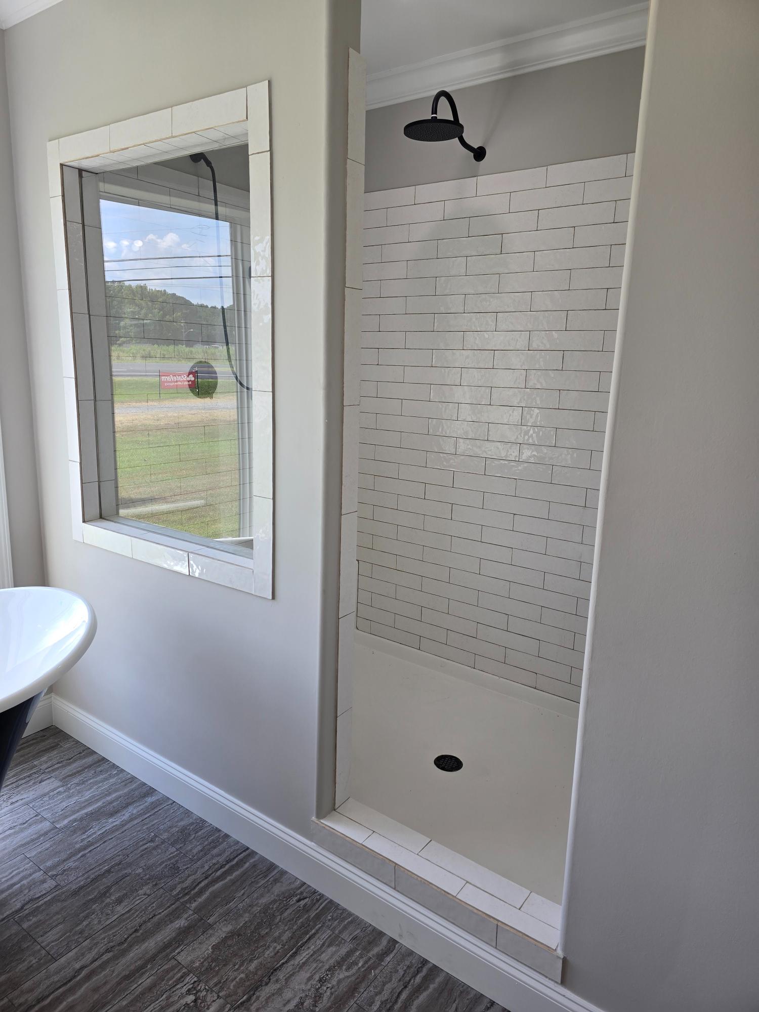 Modern bathroom with a narrow tiled shower featuring a matte black showerhead. Large window offers natural light; wood-style flooring enhances elegance.