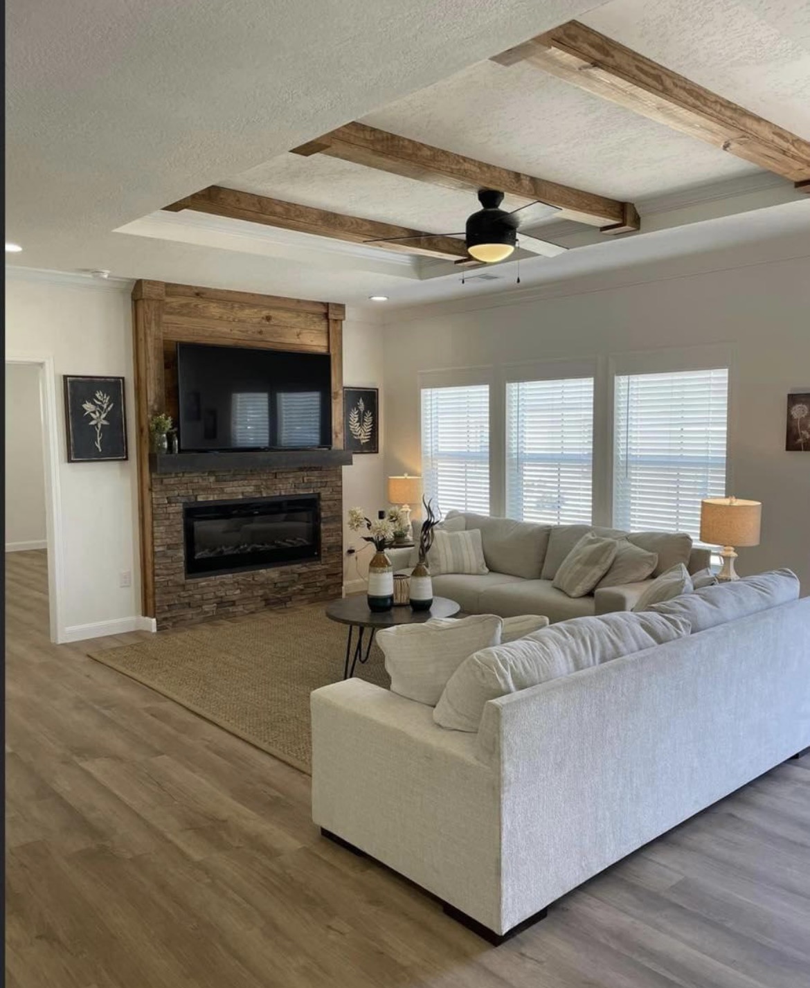 A cozy living room features a large beige sectional sofa, a stone fireplace with a TV above, wooden beams on the ceiling, and soft lighting from table lamps.