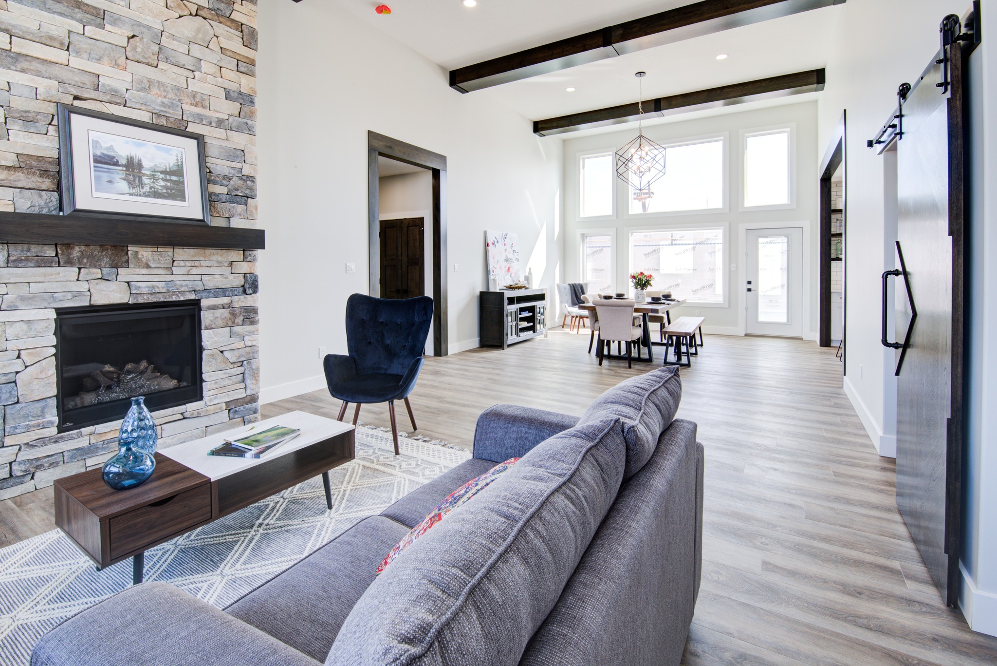 Modern living room with a stone fireplace and a cozy gray couch. Large windows and a rustic dining table create an airy, inviting atmosphere.