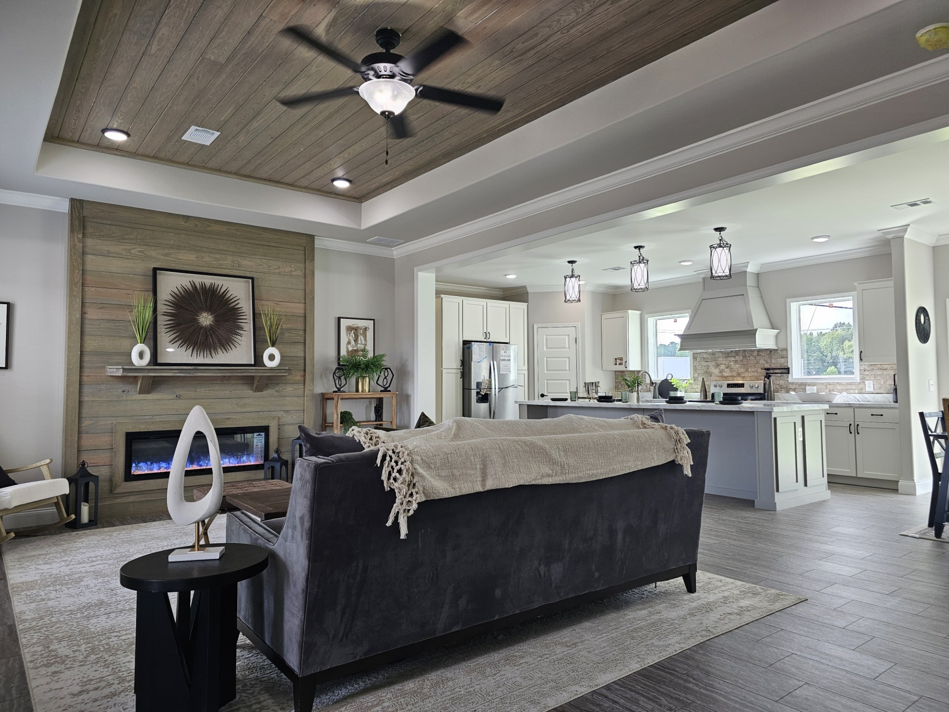 Modern living room and kitchen with wooden accents. Features a black couch, fireplace, decorative art, ceiling fan, and pendant lights. Cozy and elegant.