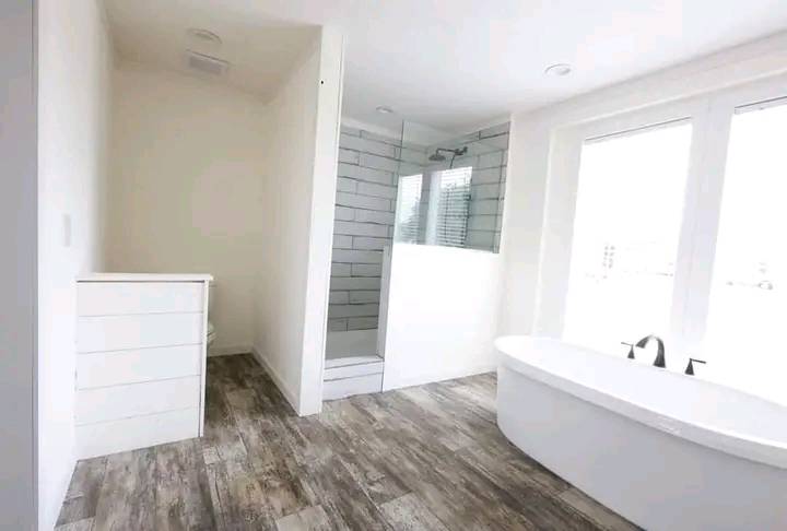Bright bathroom with wood-like tile flooring, a freestanding white bathtub, and a glass shower with white subway tiles. Large windows provide natural light.