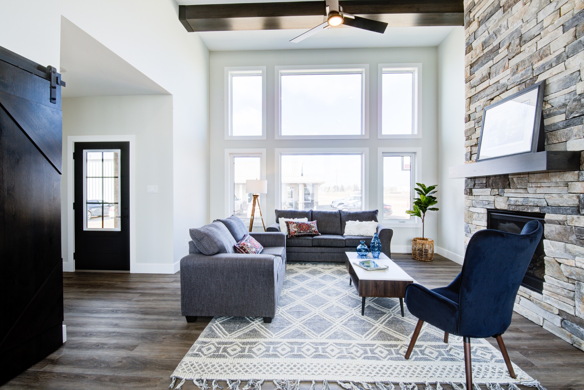 Bright, modern living room with large windows, gray sofas, a stone fireplace, wooden beams, and a cozy rug. Warm, inviting atmosphere.