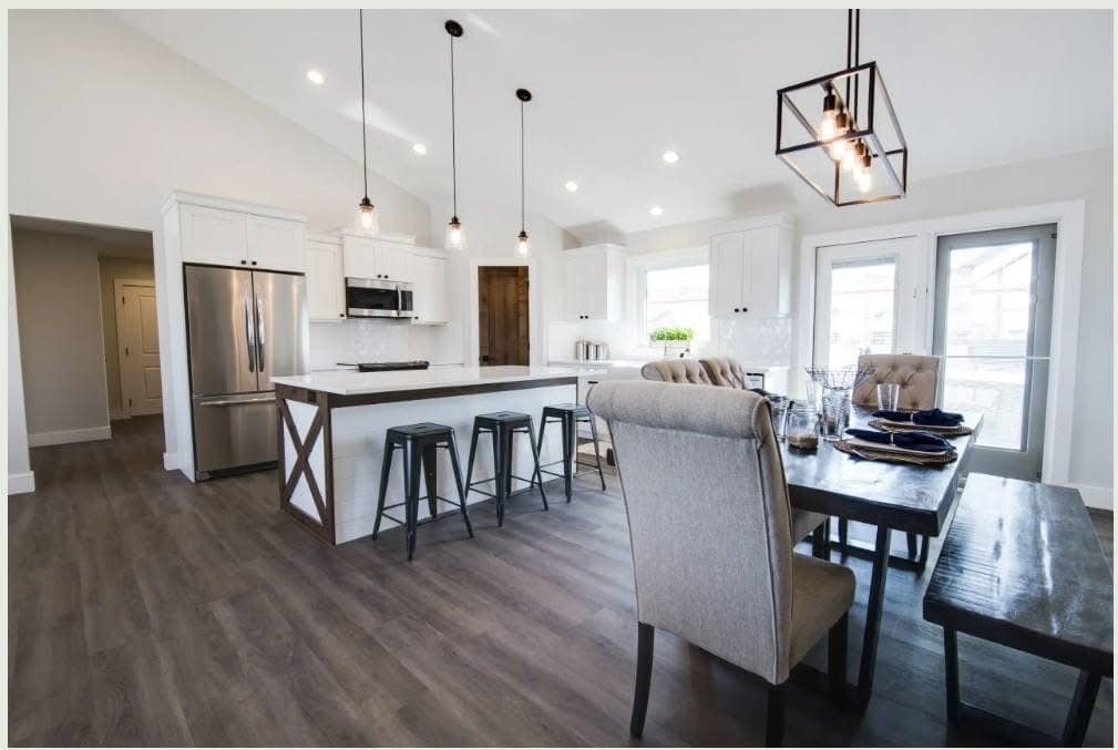 Modern kitchen and dining area with dark wood floors, white cabinets, and stainless steel appliances. Features an island with bar stools, and a dining table set for six under stylish pendant lights. Bright and spacious ambiance.