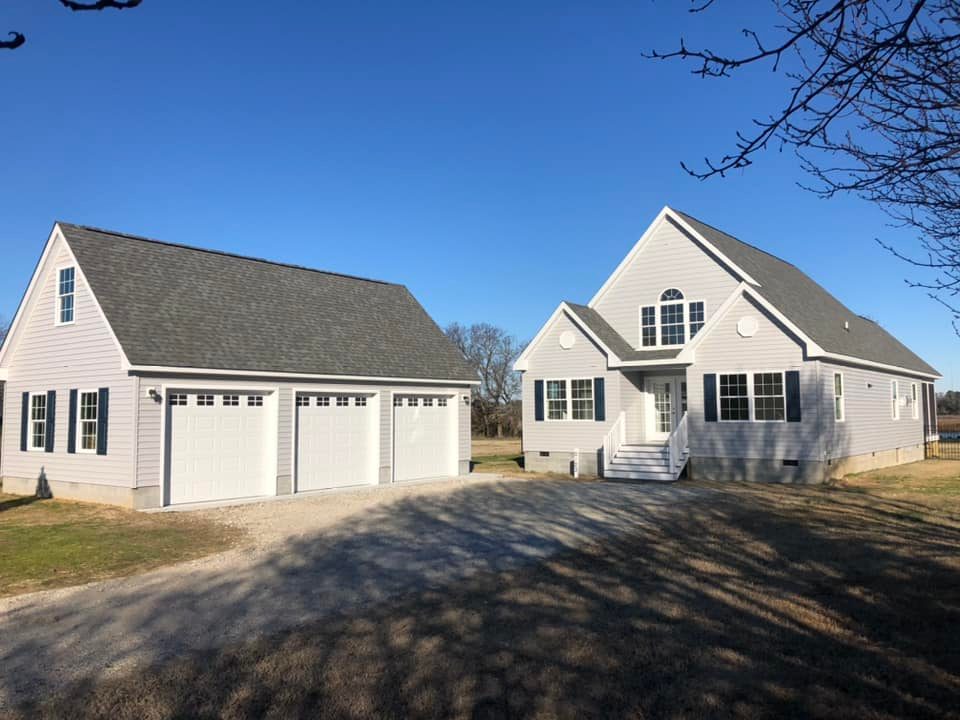 A modern, light gray two-story house with a steep roof, beside a matching three-car garage. Sunlight casts clear shadows on the lawn, under a clear blue sky.