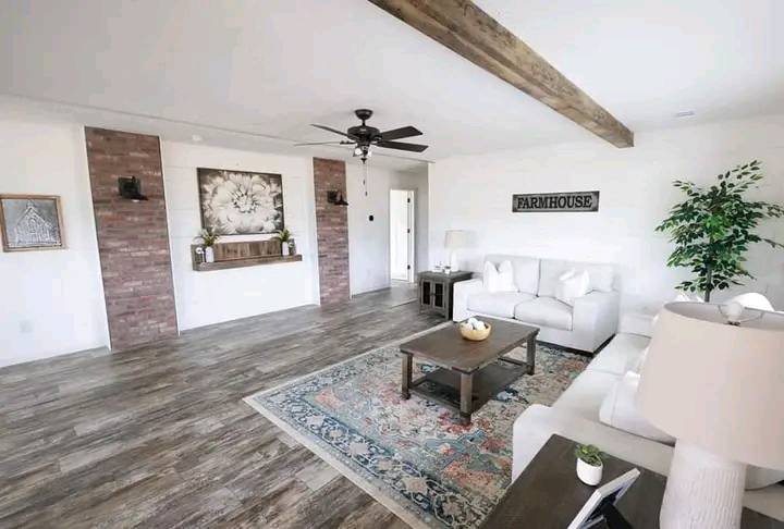 Bright living room with white sofas, a wooden coffee table on a colorful rug, brick accents, a ceiling fan, and "Farmhouse" sign. Cozy and rustic.