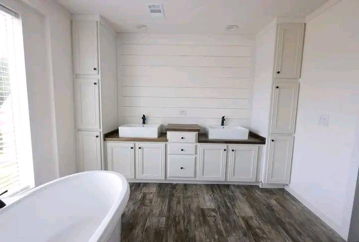 A modern bathroom features dual white sinks with black faucets set against a shiplap wall, surrounded by white cabinets. A freestanding tub is on wooden flooring, creating a serene atmosphere.