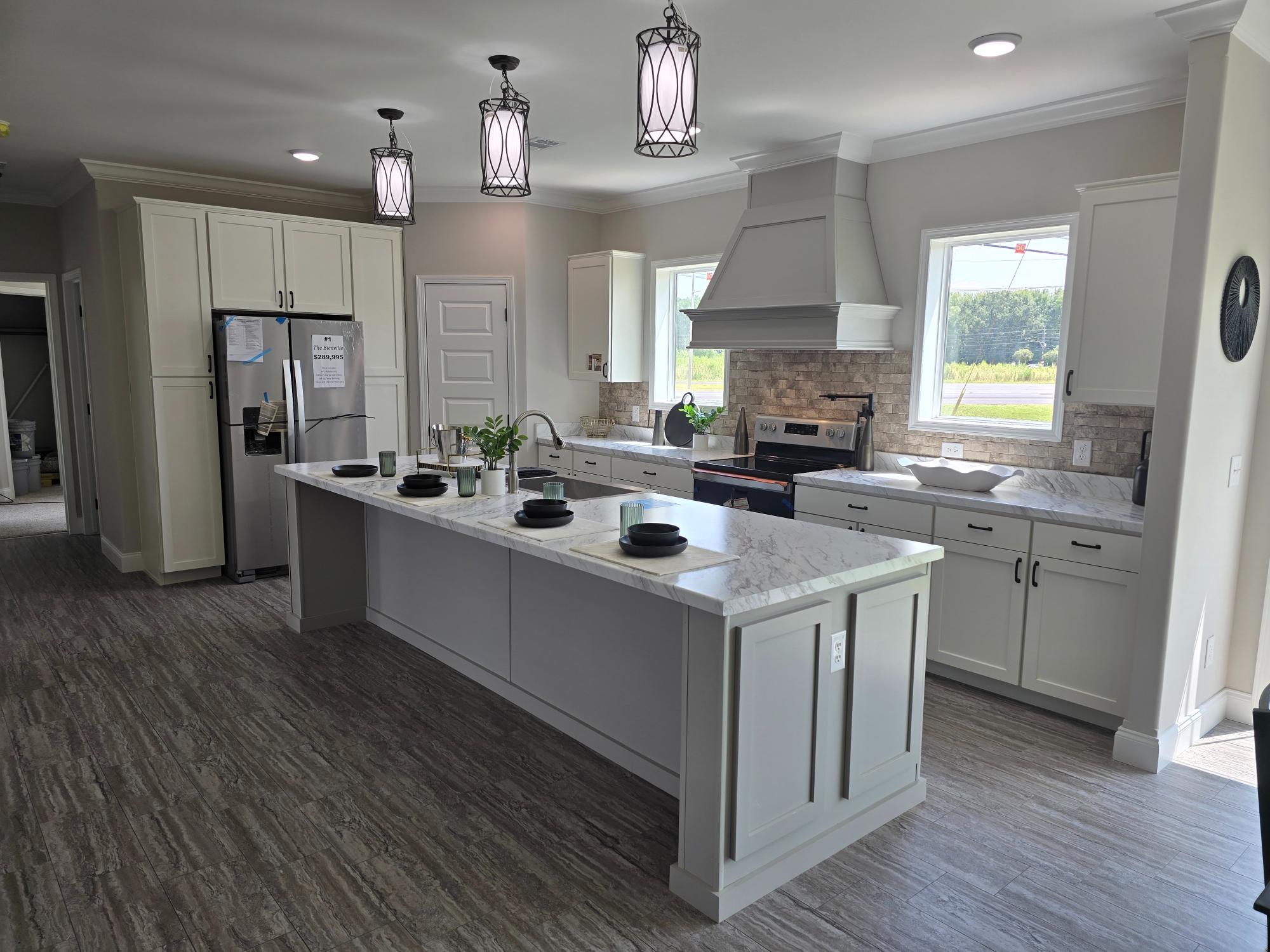 Modern kitchen with white cabinets, marble countertops, and stainless steel appliances. Central island with four black plates, pendant lights above. Airy and bright ambiance.