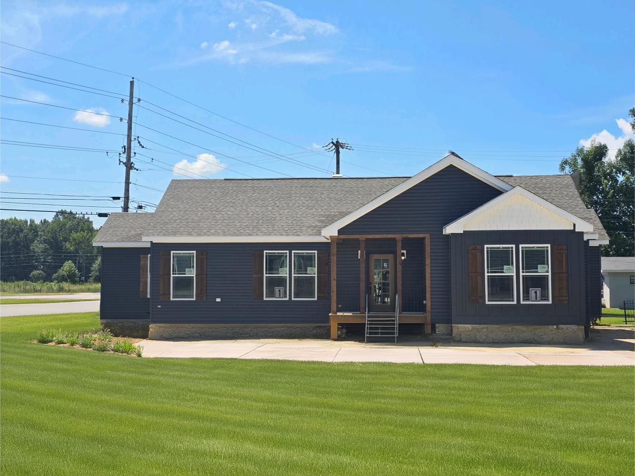 Single-story, dark-toned house with a gabled roof and wooden shutters sits on a lush green lawn under a clear blue sky, exuding a tranquil suburban vibe.