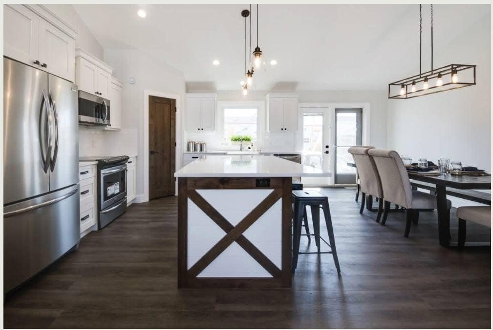 Modern kitchen with white cabinets, dark wood flooring, and a central island featuring a wooden "X" design. Light fixtures hang above, adding warmth.