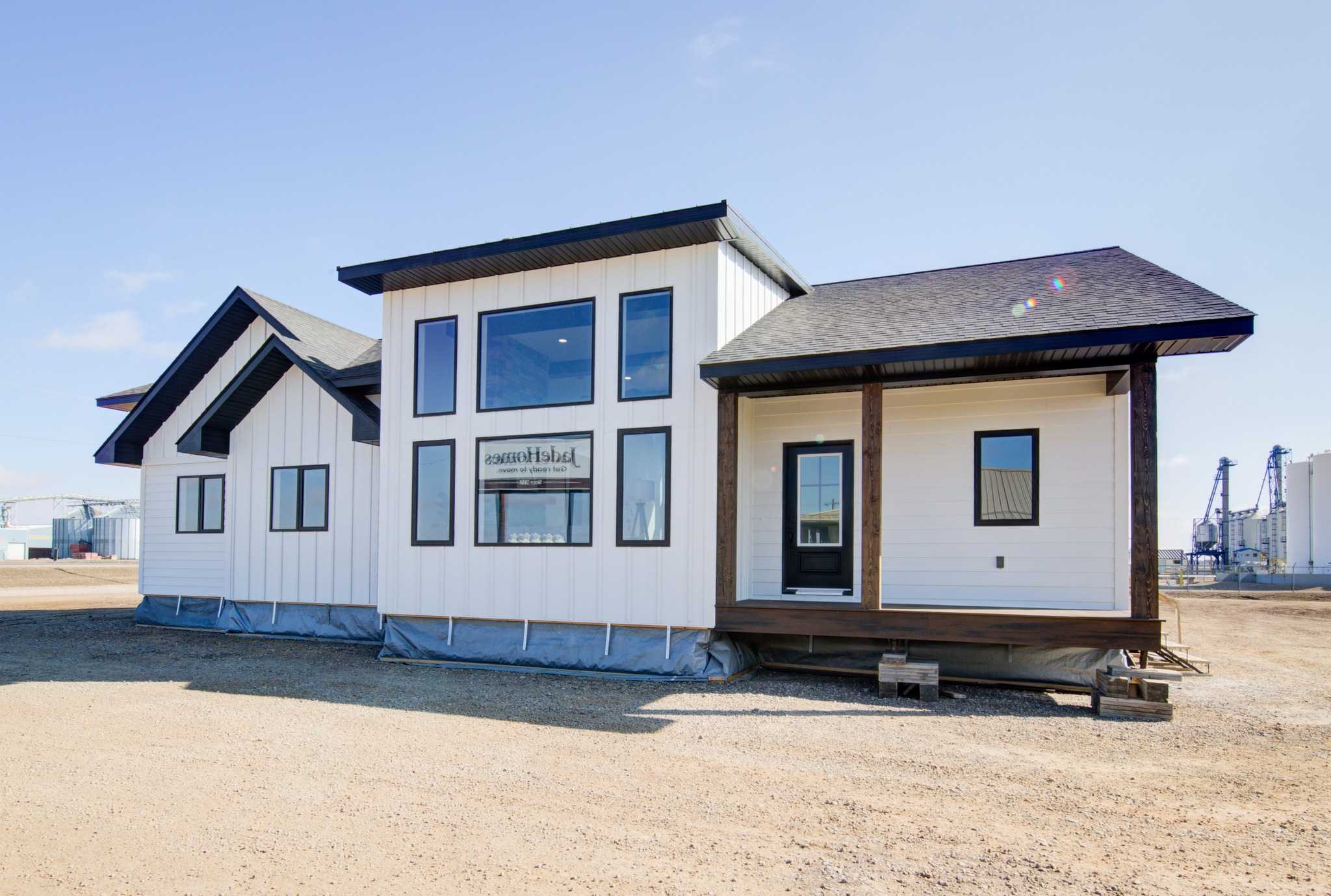 Modern white modular home with large windows and dark trim, set on a gravel lot under a clear blue sky, conveying a clean, minimalist vibe.