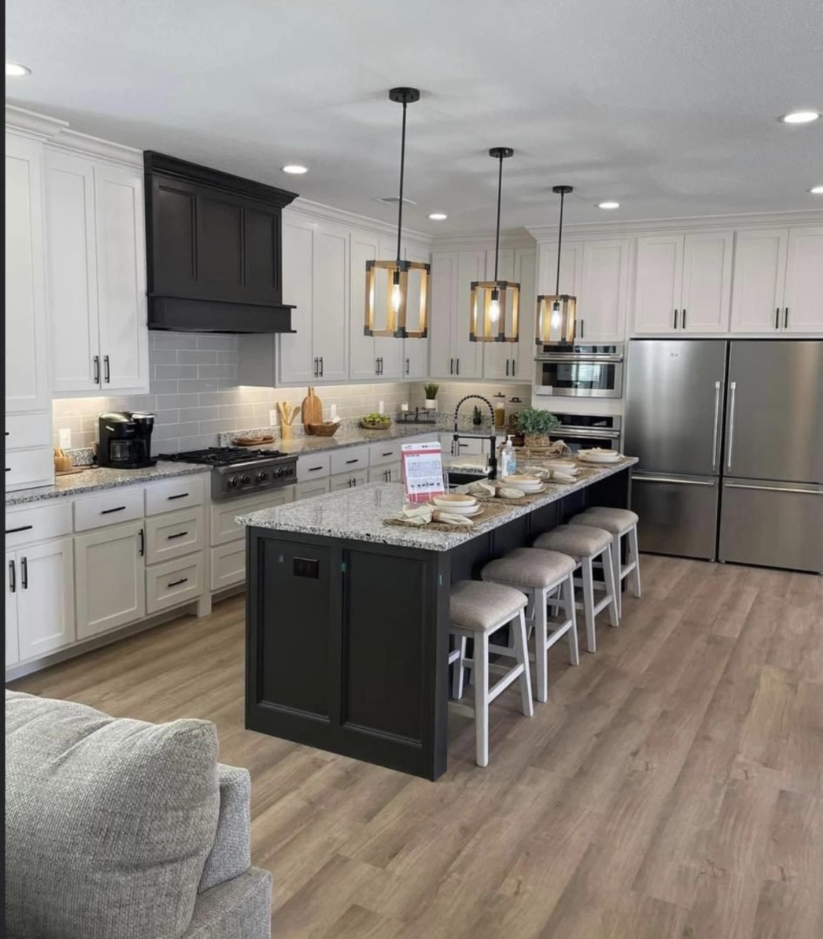 Modern kitchen with white cabinets, dark island, and granite countertops. Pendant lights hang above the island with stools. Stainless steel fridge and double oven against the wall create a sleek look. Warm, inviting atmosphere.