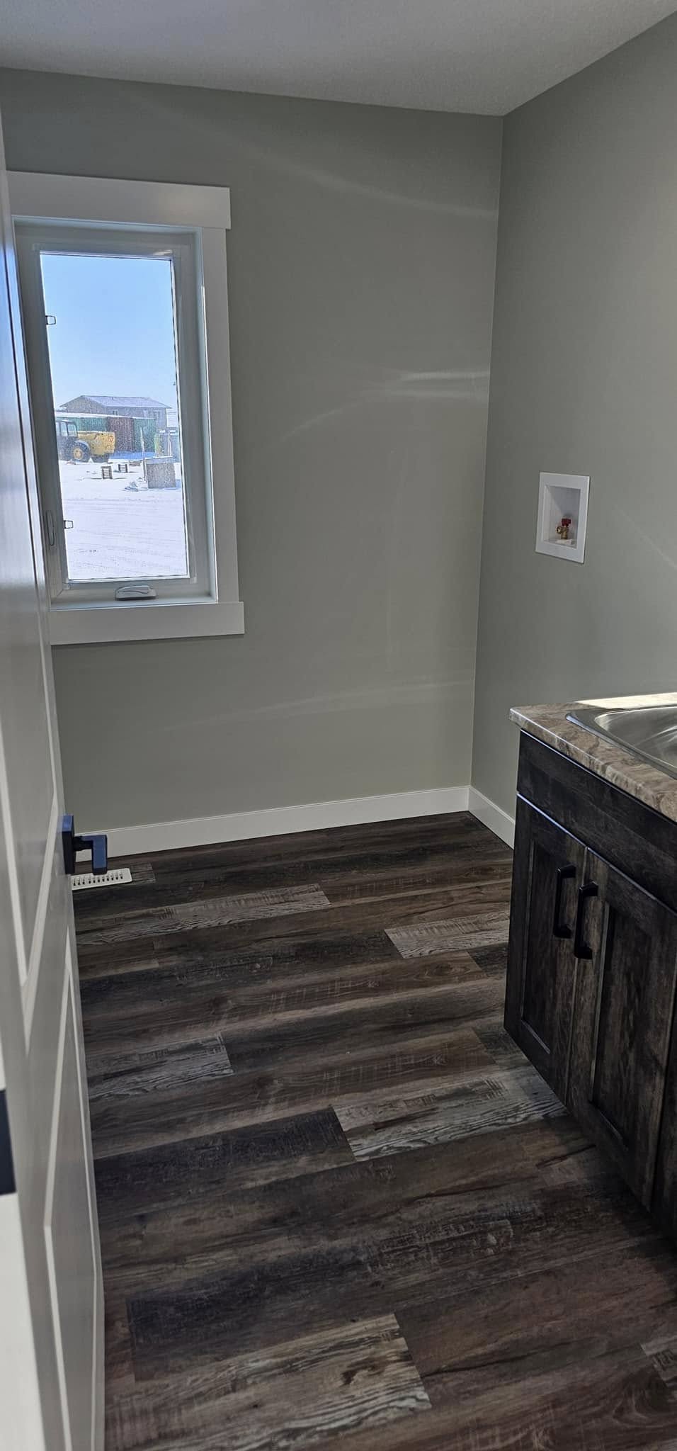A small, minimalist laundry room with gray walls and dark wood flooring. It features a single window, a wooden cabinet with a marble countertop, and a stainless steel sink. The atmosphere is clean and modern.