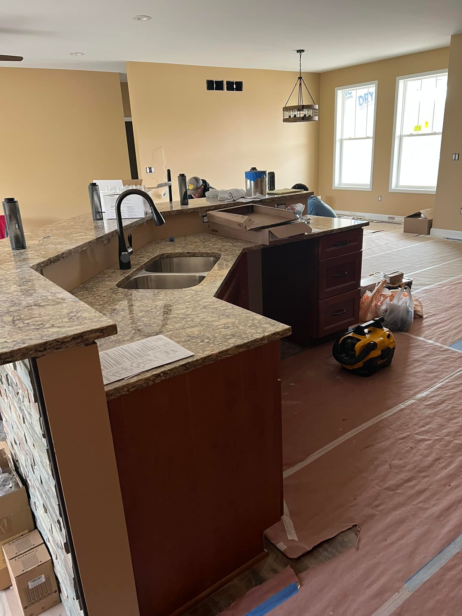 Kitchen under renovation with brown cabinets, granite countertops, and a central island sink. Tools, materials, and a vacuum are scattered on the floor.