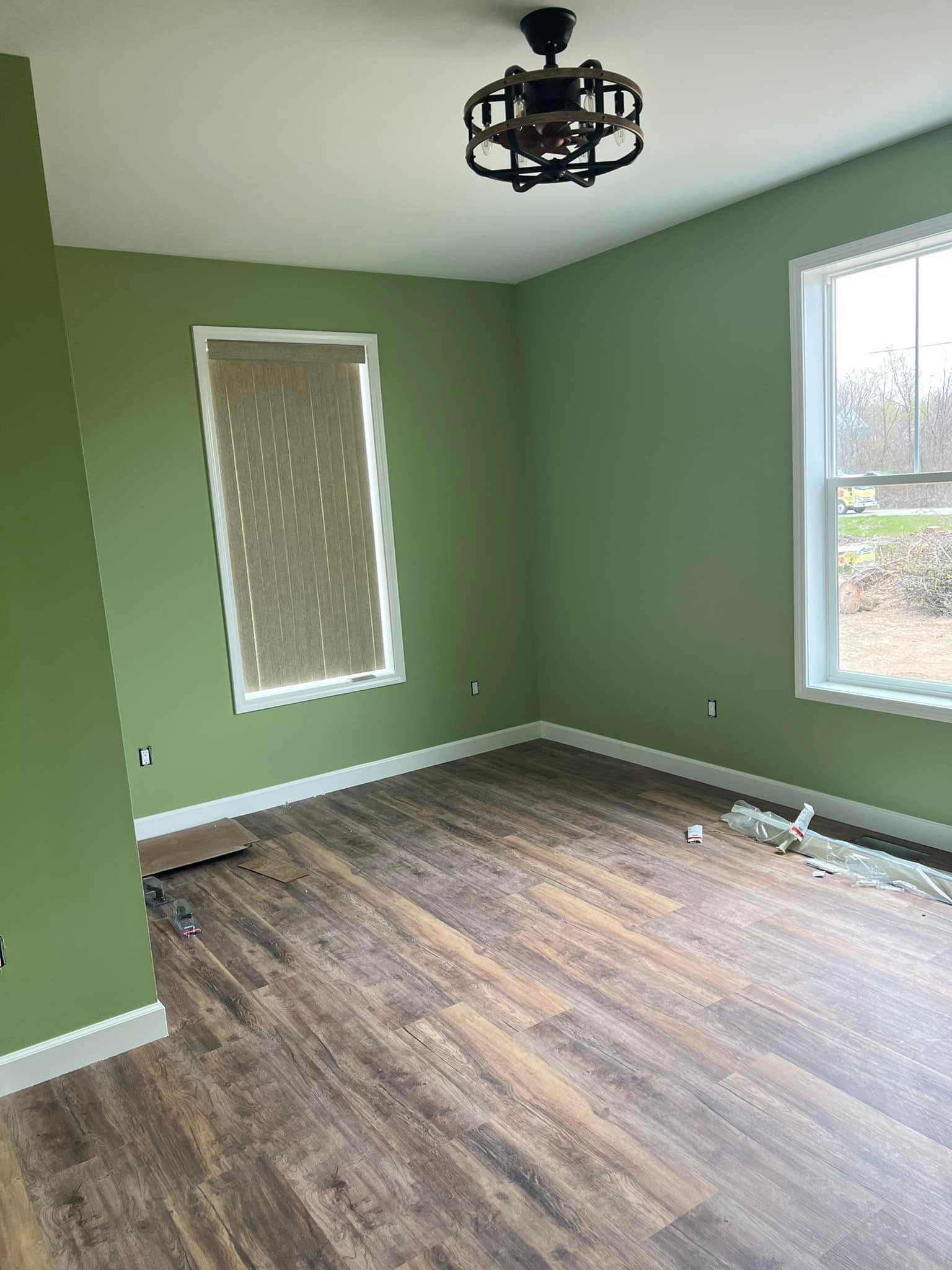 A freshly painted room with green walls, dark wood flooring, and a modern ceiling light. Two windows allow natural light, adding a calm atmosphere.