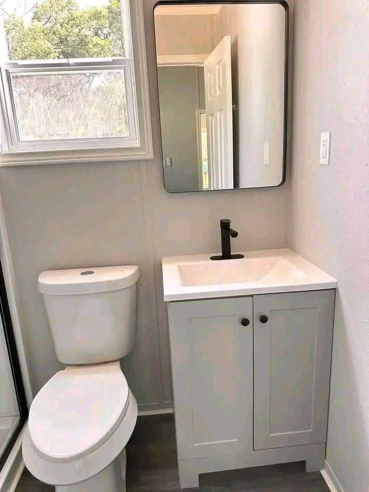 A small, modern bathroom with a white toilet and a vanity with a black faucet. A rectangular mirror hangs above. Natural light comes through a window.