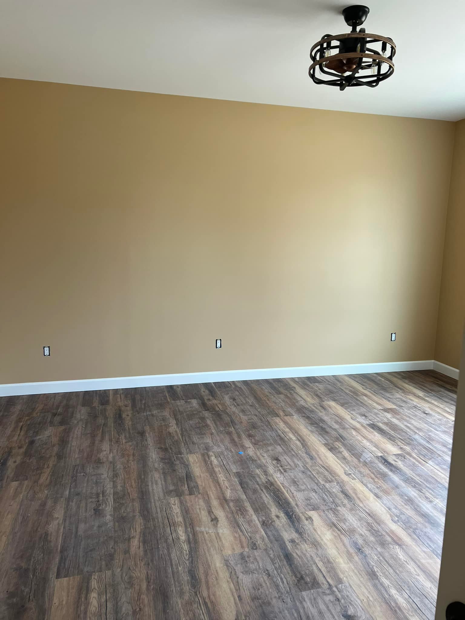 Empty room with beige walls, dark wood laminate flooring, and a modern black ceiling light. The atmosphere is clean and minimalist.