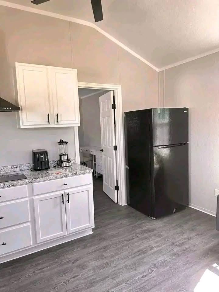 Bright kitchen with gray flooring, white cabinets, and black handles. A black fridge stands next to an open door, revealing a glimpse of another room.