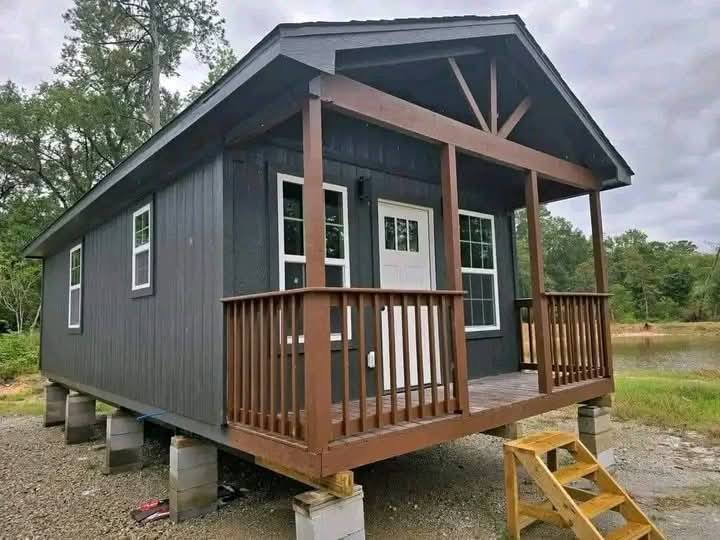 Small dark gray cabin on stilts with a porch, wooden railings, and a white door. Surrounded by trees and a pond under a cloudy sky. Rustic and cozy feel.