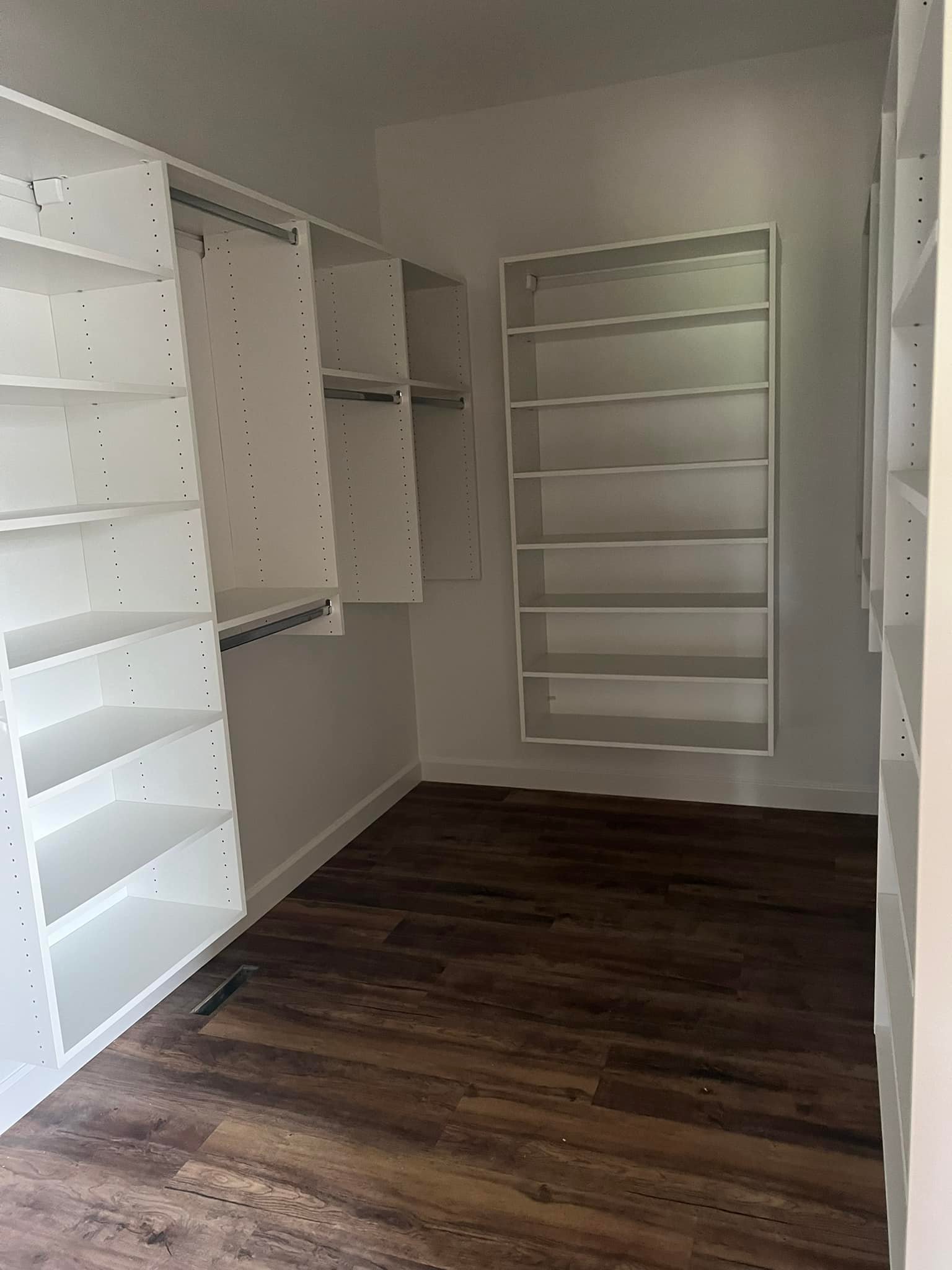 Empty walk-in closet with white shelves and hanging rods on walls. The space has a warm, brown hardwood floor, creating a clean, organized feel.