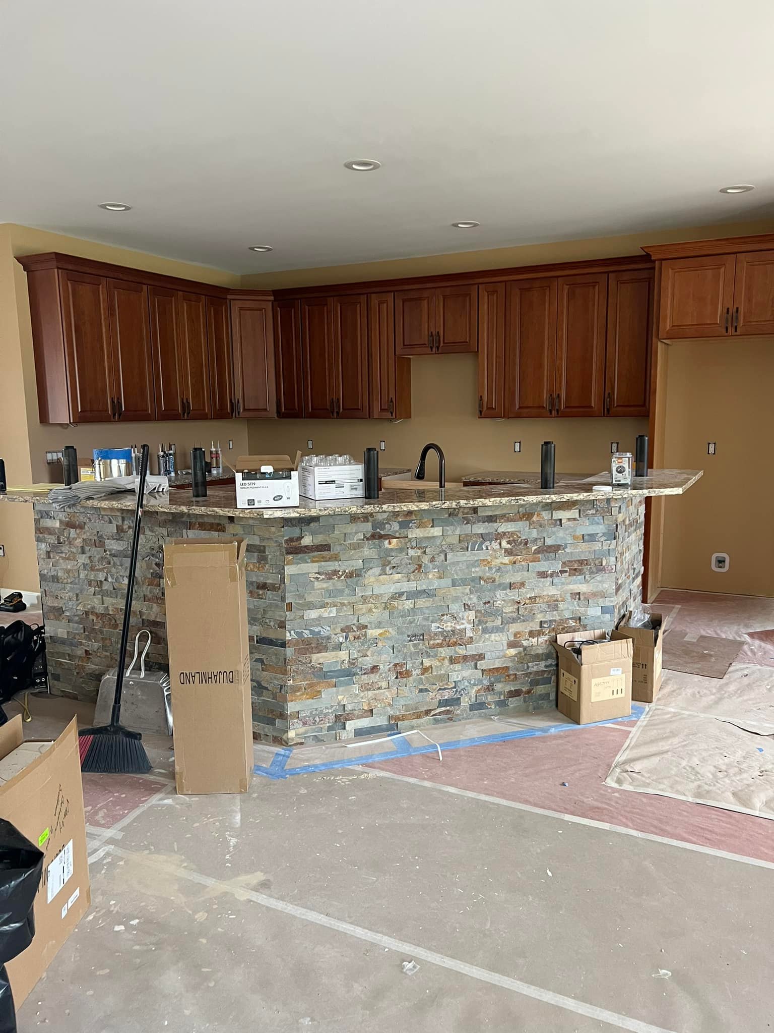 Kitchen under renovation with stone island, granite countertop, and brown cabinets. Boxes, tools, and a broom scattered; a work in progress.