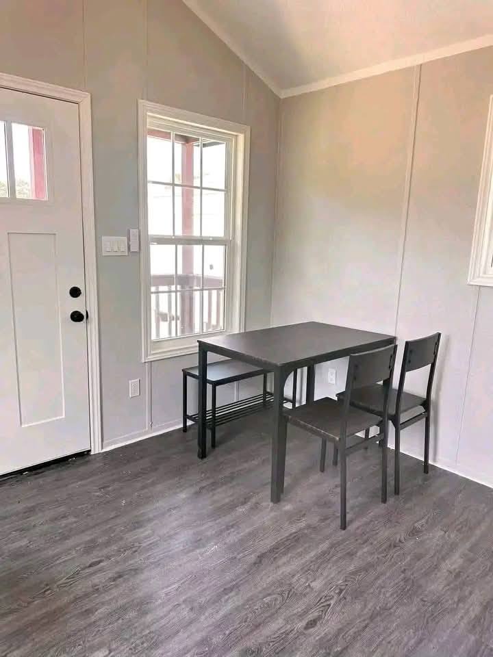 A minimalist room with gray walls features a small dark gray table, two chairs, and a bench. Natural light enters through a window, creating a simple, calm ambiance.