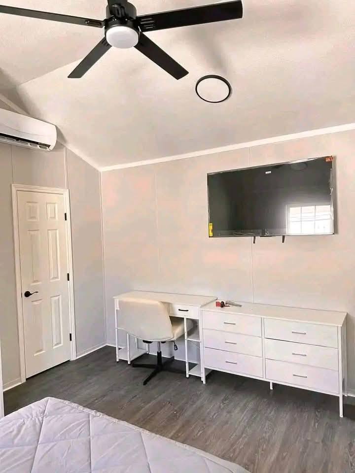 Bright and minimalist bedroom featuring a ceiling fan, wall-mounted TV, white desk and chair, dresser, and wood flooring, creating a clean and modern ambiance.