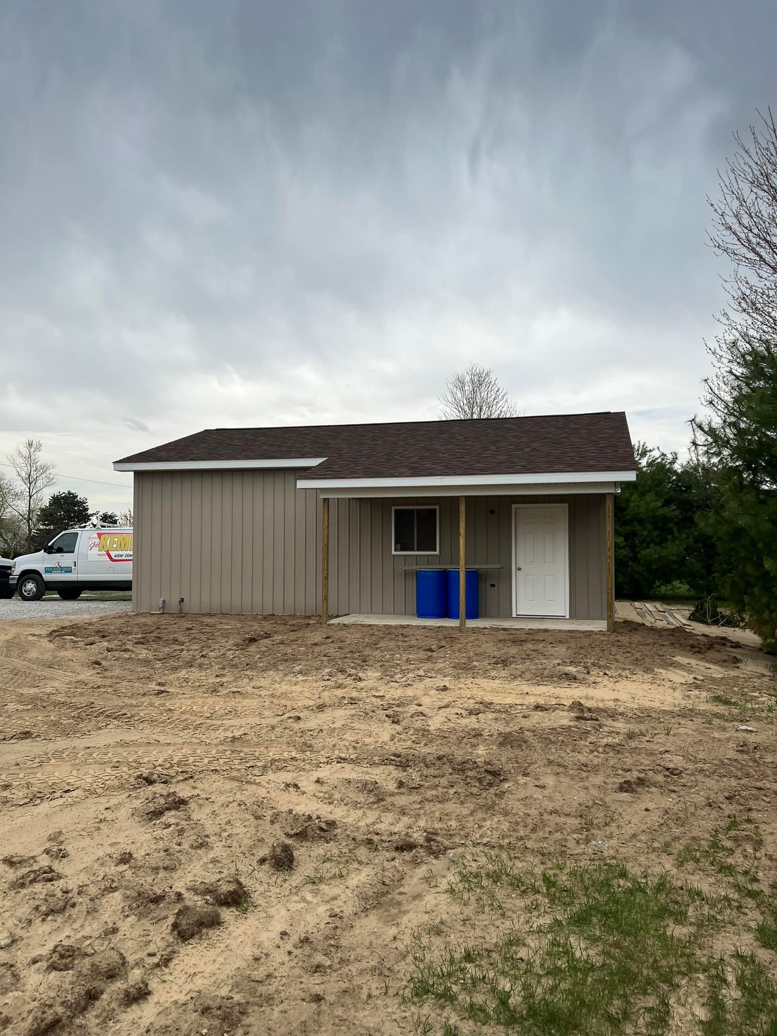 Small beige building with white door and overhang, set on a bare dirt patch. Overcast sky above. A parked van is partially visible on the left.