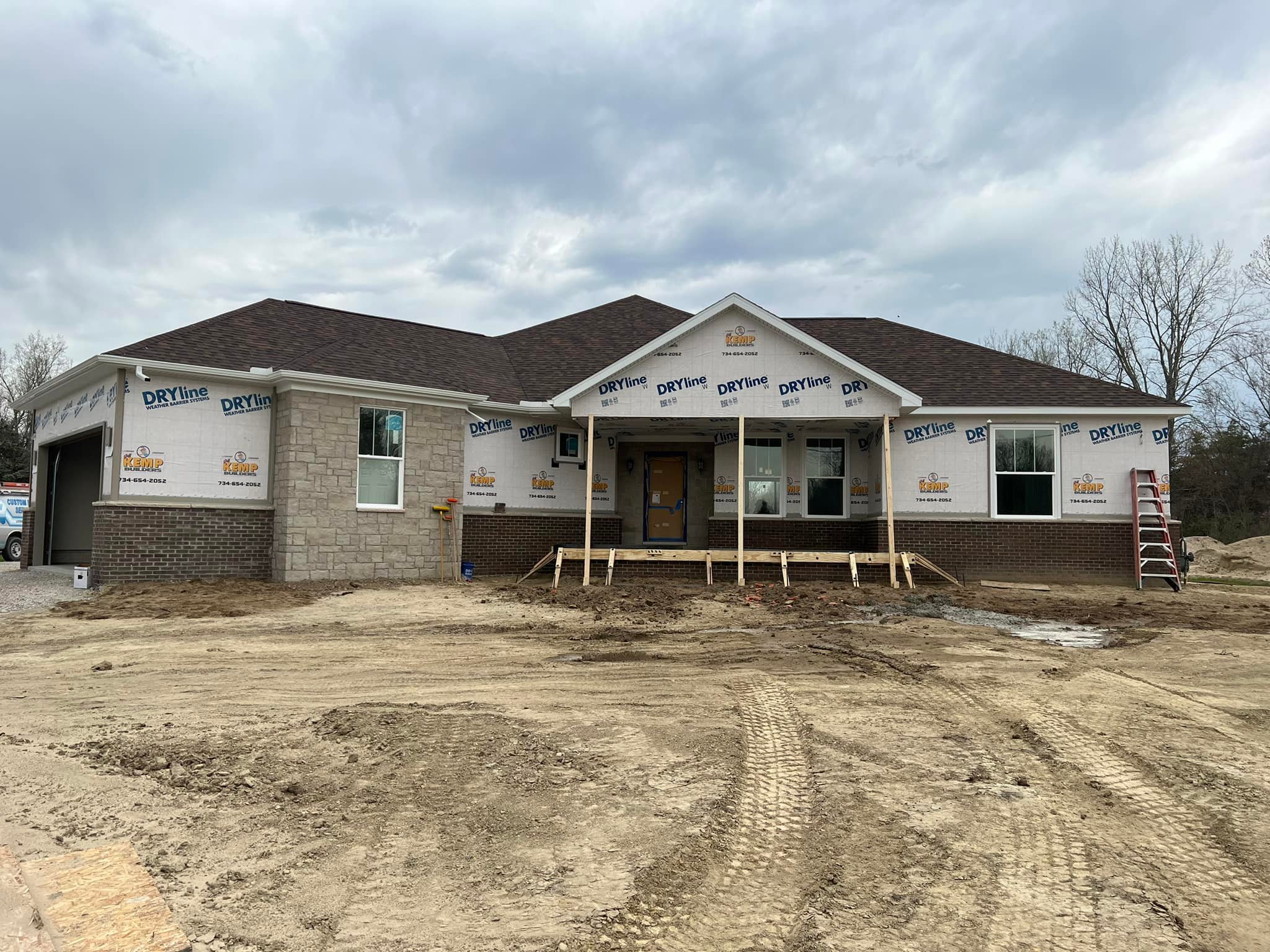 Single-story house under construction with unfinished front steps. The exterior features partial brickwork and visible building wrap. Cloudy sky.