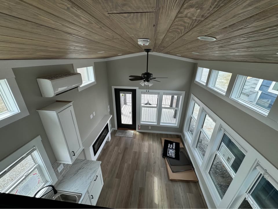 Alt text: Modern tiny home interior with wood ceiling and floor, white cabinetry, a ceiling fan, and ample windows, creating a bright, airy atmosphere.